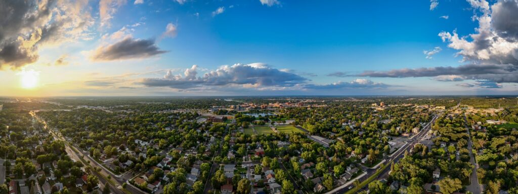 Panoramic view of Saint Cloud, MN, showcasing University Drive South, Lake George, City Hall, most of downtown, the Mississippi River, and a stunning sunset in the background.